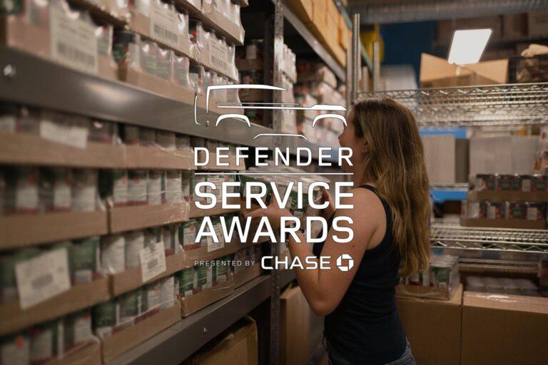 A woman stands in a storage room with shelves of boxes and cans. The text overlay reads “Defender Service Awards presented by Chase” with a vehicle outline.
