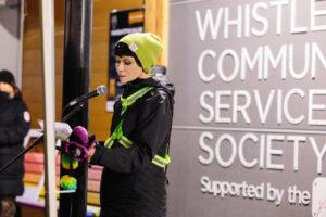 A person in a green hat and reflective jacket speaks at a microphone in front of a Whistler Community Services Society sign.