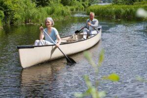Two older adults paddle a beige canoe on a calm, narrow river surrounded by greenery and tall grass.