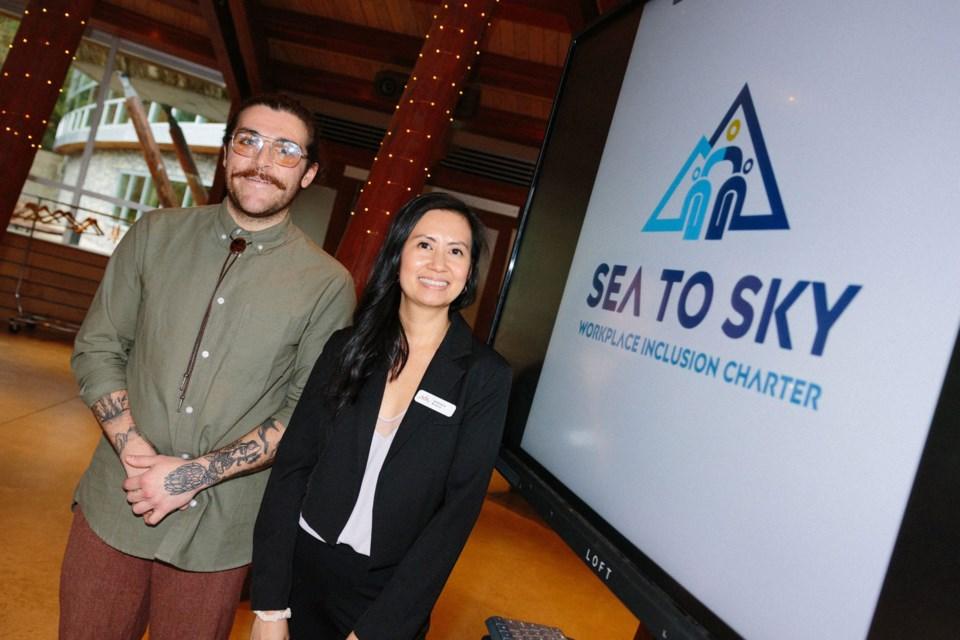 Two people stand indoors next to a screen displaying the Sea to Sky Workplace Inclusion Charter logo at an event.